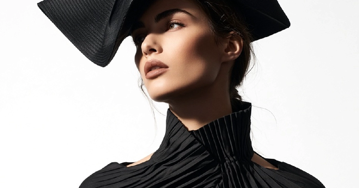 Model in black pleated dress and architectural hat on white seamless